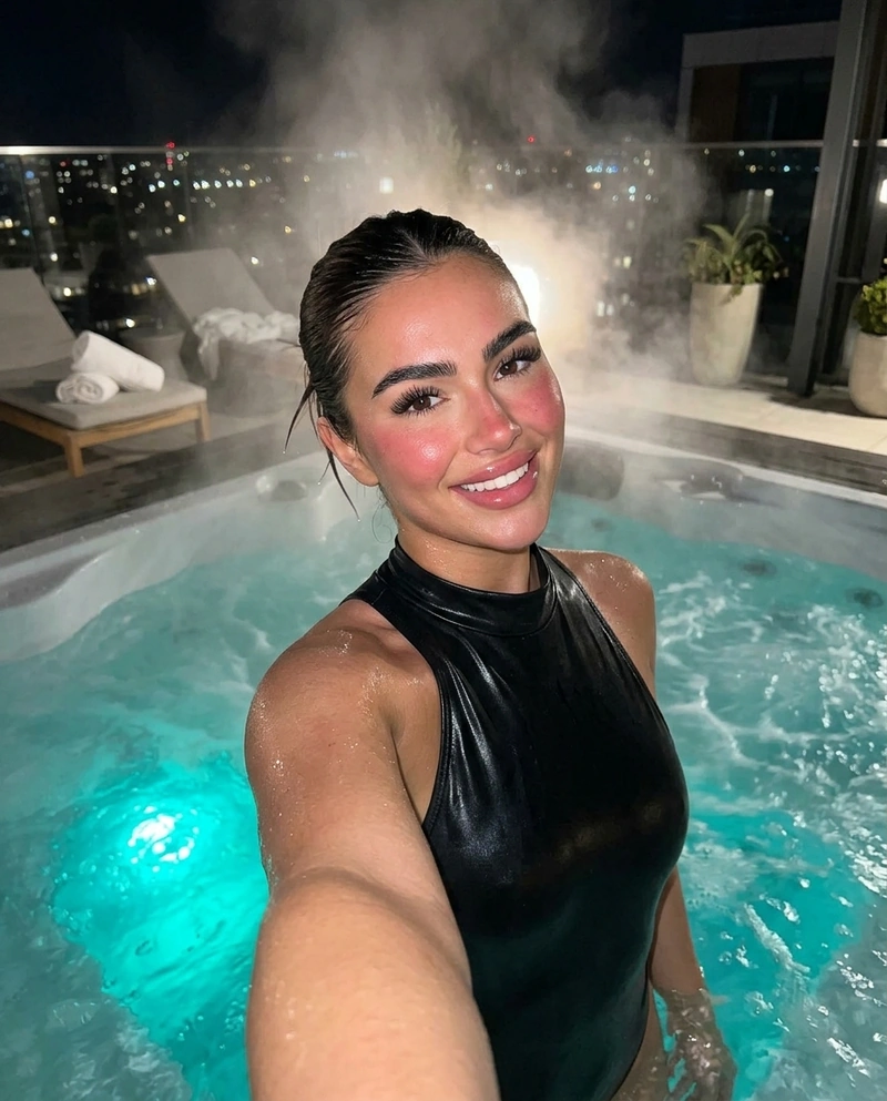 AI-generated: Front-facing selfie taken by a beautiful young woman standing in a steaming rooftop hot tub at night, one arm extended holding the camera. S