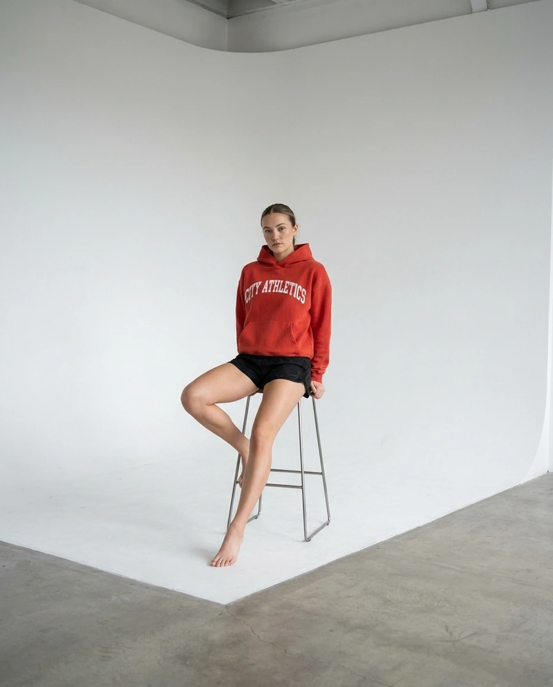 AI-generated: A beautiful 22-year-old female model wearing the @baba hoodie and short, tight black polyester running shorts, sitting on a high chair. Stud