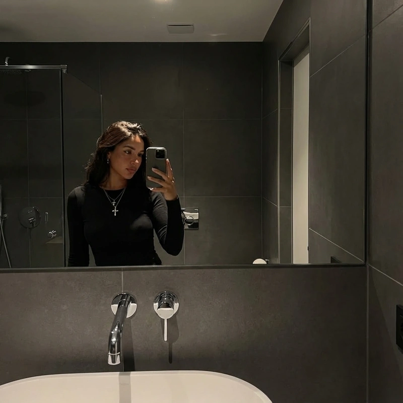 AI-generated: {
  "prompt": "A young woman with long chestnut-brown hair takes a mirror selfie in a modern grey-tiled bathroom. She wears a black fitted l