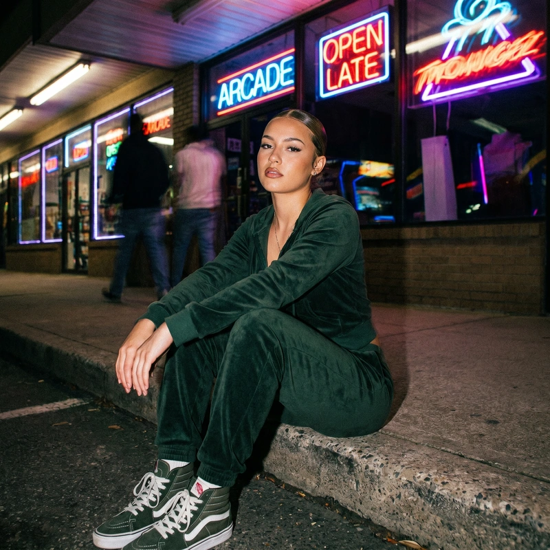 AI-generated: Young woman in forest green velour tracksuit seated on concrete curb, confident direct gaze, sleek pulled-back hair, high-top sneakers. nigh