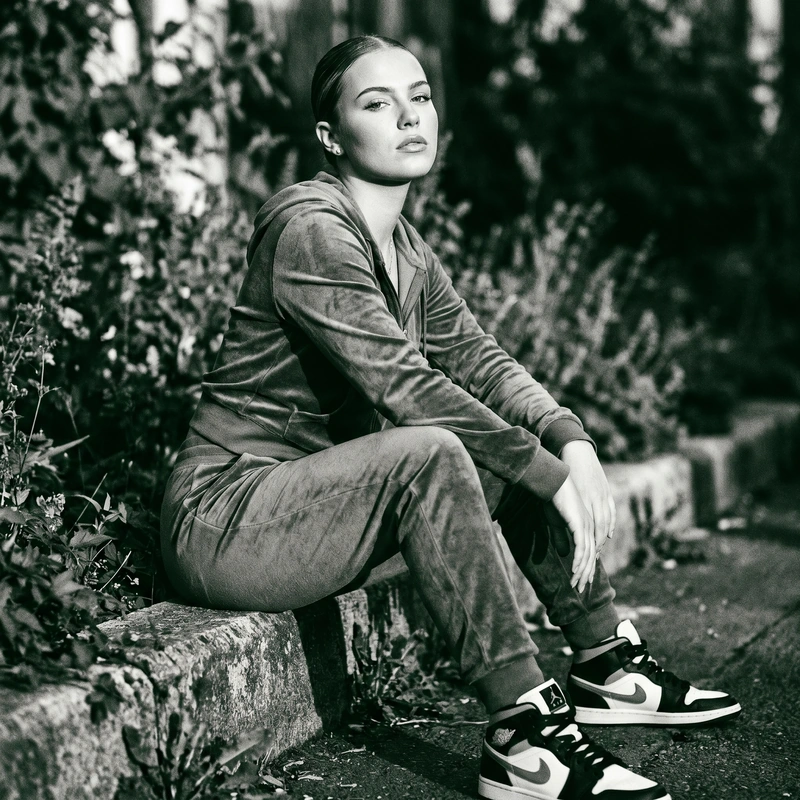 AI-generated: Young woman in forest green velour tracksuit seated on concrete curb, confident direct gaze, sleek pulled-back hair, black and white high-to